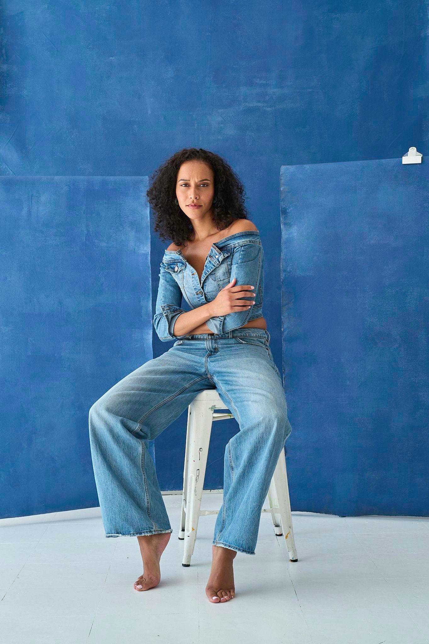 Woman with curly hair in a denim top and wide-leg jeans, seated barefoot on a white stool against blue backdrops