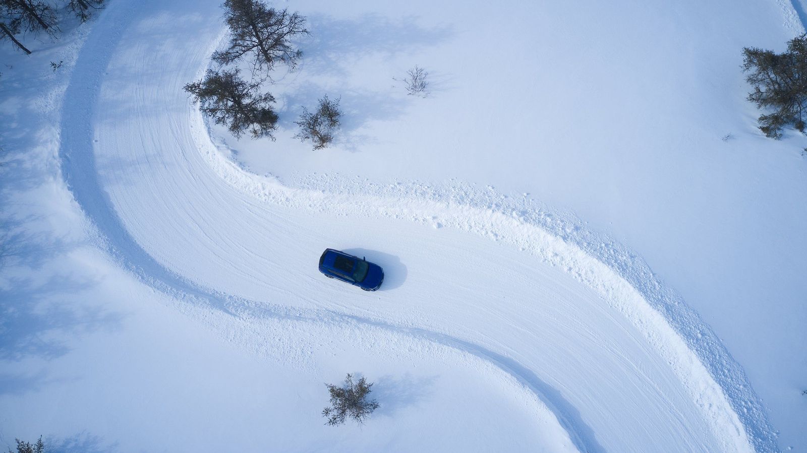 Aerial view of a blue car on a winding snow-covered road, tire tracks curving between scattered small trees