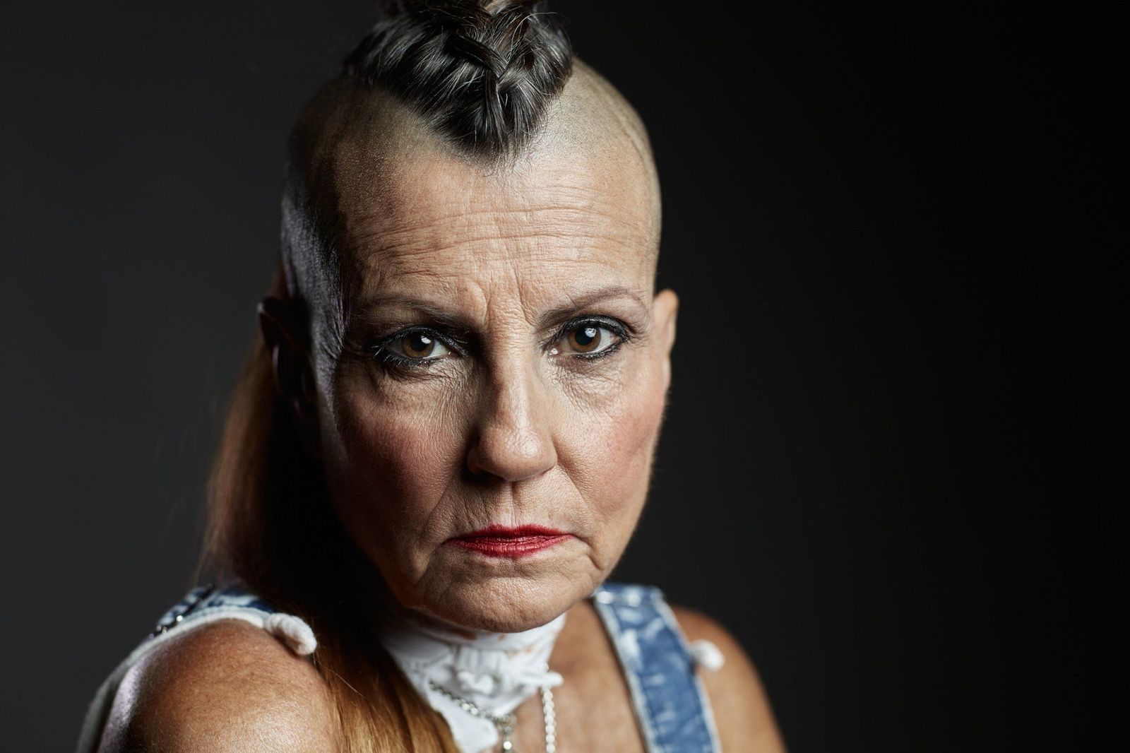 Studio portrait on black of an older woman with a braided mohawk, red lipstick and a steady, direct stare