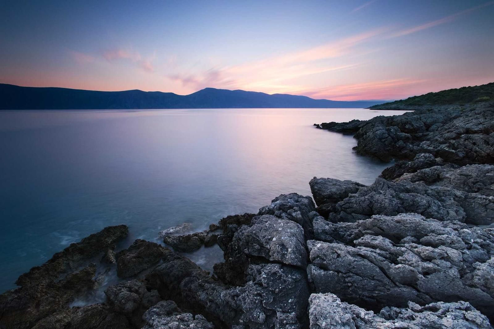 Serene twilight seascape with rocky coastline, calm turquoise waters, and distant mountains at dusk