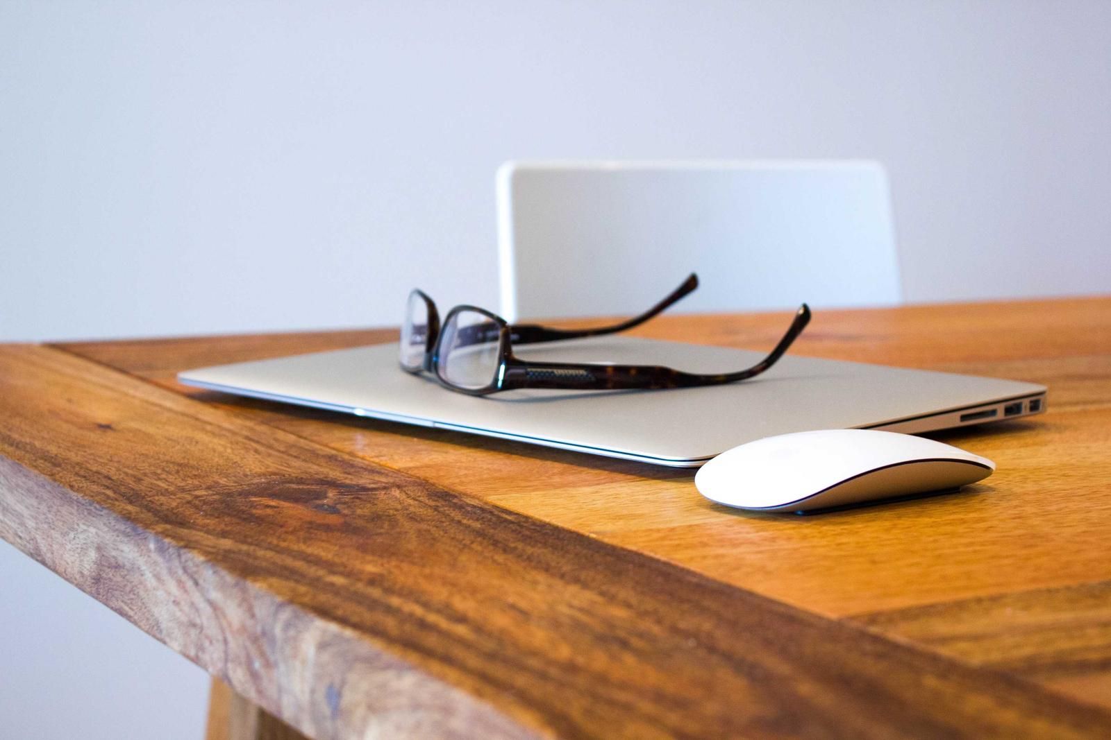 Minimalist workspace setup on wooden desk with silver laptop, wireless mouse, eyeglasses, and monitor against white background